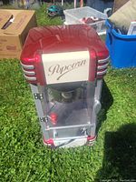 Red and silver tabletop popcorn maker on grass, showing glass panels and retro 'Popcorn' label on front.