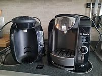 Two Bosch Tassimo coffee makers side by side on countertop. Both black color, clearly showing control knobs, pod compartments, and drip trays.