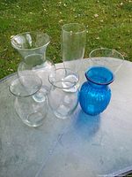 Photo showing six glass vases on an outdoor surface, including five clear glass vases and one blue ribbed vase.