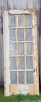 Vintage wooden door with 15 rectangular glass panels, heavily peeling paint, standing upright against a wooden wall.