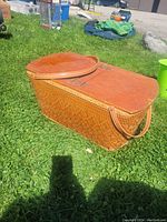 Photo showing a large rectangular vintage picnic basket in woven material with two hinged wooden lids and a handle, photographed outdoors on grass.