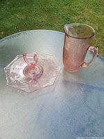 Photo of an octagonal pink glass plate with a looped handle in the center and a tall matching pink glass pitcher with etched floral design, placed on a glass table outside.