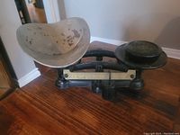 Photo of a vintage mechanical balance scale showing a curved metal weighing plate on the left and a flat circular plate on the right, placed on a wooden table.