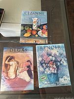 Three hardcover art books featuring Cézanne, Degas, and Renoir, arranged on a glass table.