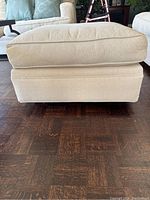 Front view of the beige pinstripe fabric ottoman showing its rectangular shape, cushioned top and base, and wheel visibility beneath.