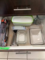 Photo of assorted rectangular food containers with green lids, clear food storage dishes, and metal box grater in drawer