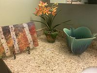 Photo showing decorative plate with birch design, faux flower arrangement in green pot, and green leaf-shaped glass vase on countertop.