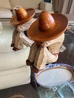 Pair of hand carved wooden figures wearing large sombreros, viewed from side and front angles on glass table.