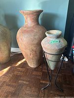 Two ceramic pots side by side on wooden floor, taller reddish-brown pot and smaller greenish-gray pot on black stand visible.