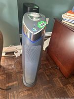 Photo of GermGuardian tower air purifier standing upright on floor, black and silver color with blue accent light, control buttons on top