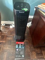 Full front view of the black Honeywell tower air purifier on wooden floor beside furniture, showing control panel and additional K filter box placed in front of it.
