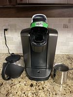 Photo shows a Keurig coffee maker with a glass teapot on the left and a stainless steel milk frothing pitcher on the right, all placed on a kitchen granite countertop.