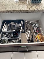 Top view of kitchen drawer showing organized trays with assorted forks, spoons, knives, and several kitchen utensils including white handle spreaders and wine openers.