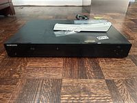 Front view of the Samsung Blu-ray player with remote on top, showing the black glossy finish and front panel details.