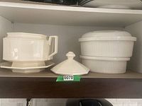 Photo showing white octagonal ceramic stew server with lid, matching serving plate, and two ribbed round casserole dishes with lids stored in a cabinet shelf.