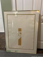 Full front view of framed artwork showing embossed abstract design with gold accents, glass and gold frame, standing against white door.