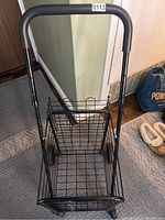 Black metal shopping cart with wire basket, padded handle, and wheels for mobility, positioned on a carpeted floor indoors.