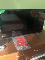 Front view of TCL TV on wooden cabinet with manuals and cables in plastic bag in front.