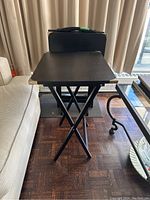 Front view of a black wooden TV tray with brass corners on folding legs, set up beside a sofa.