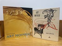 Photo of two Art Nouveau hardcover books upright on wooden surface, showing front covers with different artistic styles related to Art Nouveau.