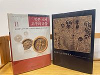 Two hardcover books standing on a wooden table, one titled 'Arts of Korea' with an ornate gold and black cover, the other with Korean text and images of ancient artifacts.