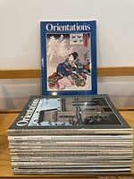 Stack of 10 Orientations magazines with predominately photographic covers, several issues visible, standing vertically against wall.