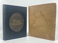 Two vintage travel books standing upright side by side, showing front covers. One black hardcover with oval emblem, one brown cloth cover with embossed patterns, vintage from mid 1800s.