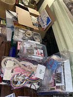 Wide shot of multiple craft supplies including yarn, stitch painter, buttons, knit calculator, rotary blades, and assorted tools organized in boxes and bags on floor.