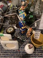 Overview of lot showing decanters, figurines, thermometers, picture frames and ceramic items on fabric-covered table.