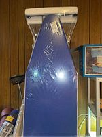 Blue ironing board with plastic wrapping and white handle at narrow end.
