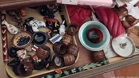 Top view of wooden napkin rings, small painted figurines, decorative ceramic plate with floral design, red twisted candle, and lidded metal dish inside a drawer.