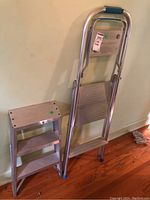 Two aluminum metal folding step stools side by side, one smaller with three steps and one taller with two steps and handle grip, showing general condition and structure.
