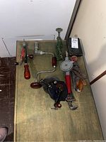 Wide shot of assorted vintage hand tools on green surface including hand drills, pipe wrench, clamp, and hand crank grinder.