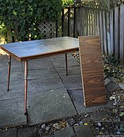 Wide view of the rectangular walnut pattern Arborite/Formica table with original leaf standing next to it outdoors on stone paving