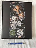 Frontal photo of the artwork titled 'Redemption' by Angelina Wrona showing a woman with three cats and bubbles, black background, with a pen for scale.