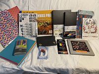 Top down photo of various art supplies including sketchbooks, canvas pad, paint by pixel kit, coloring book, markers, colored pencils, crayons, pastel oil sticks, colored card stock, and a small pouch.