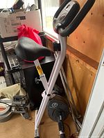 Side view of the Marcy folding stationary bike showing the black cushioned seat, pedals, and small digital display. The bike is positioned indoors near a window with boxes and other items in the background.