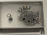 Close-up photo of Hello Kitty rhinestone brooch and small silver-tone charm in a small box with cushioning.
