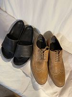 Photo showing pair of black Birkenstock slide sandals next to tan brown leather wingtip dress shoes on white surface.