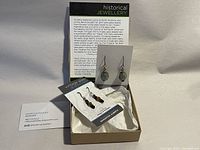 Two pairs of earrings and a pendant displayed in a box with a card describing historical glass jewelry and artist details.