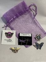 Photo of enamel black and white butterfly brooch, enamel bee brooch, vintage Mexican alpaca silver and abalone shell turtle brooch, blue glittery butterfly pin, dragonfly earrings packaged with price tags on a purple sheer drawstring pouch.
