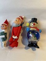 Photo showing two small felt knee hugger elf figurines and a vintage plastic snowman figure with a clear belly depicting a snowman inside. All three items have a vintage look consistent with the 1960s era.