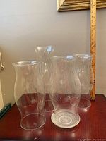 Four tall clear glass chimneys grouped together for size comparison with ruler at back