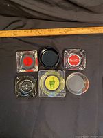 Top view of all six ashtrays arranged with a ruler for scale, showing various logo designs and clear to tinted glass styles.