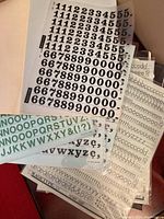 Multiple vintage dry rub-on letter and number sheets in black and green, spread out on surface for view. Sheets include numeric sheets and alphabet letter sheets.