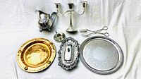 Full view of all items on white background including silver creamer jug, candelabra candlestick, salt cellar, tongs, butter dish, silver platter, and hammered copper plate
