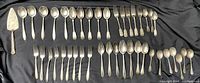 Full collection of silverplate cutlery laid out on black cloth, showing the variety of spoons, forks, and a pie server, all with vintage wear.