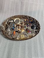 Top view of vintage ornate oval mirror tray filled with assorted jewelry items including bracelets, necklaces, earrings, and rings.