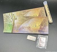 Two KaratPal CashGold envelopes, a plastic tube of gold flakes, a generic silver bar and a Northwest Territorial Mint bar on black background