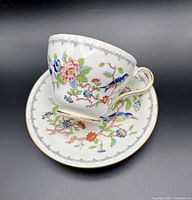 Tea cup and saucer from a slight angled top view showing floral and bird decorative pattern with gold trim.